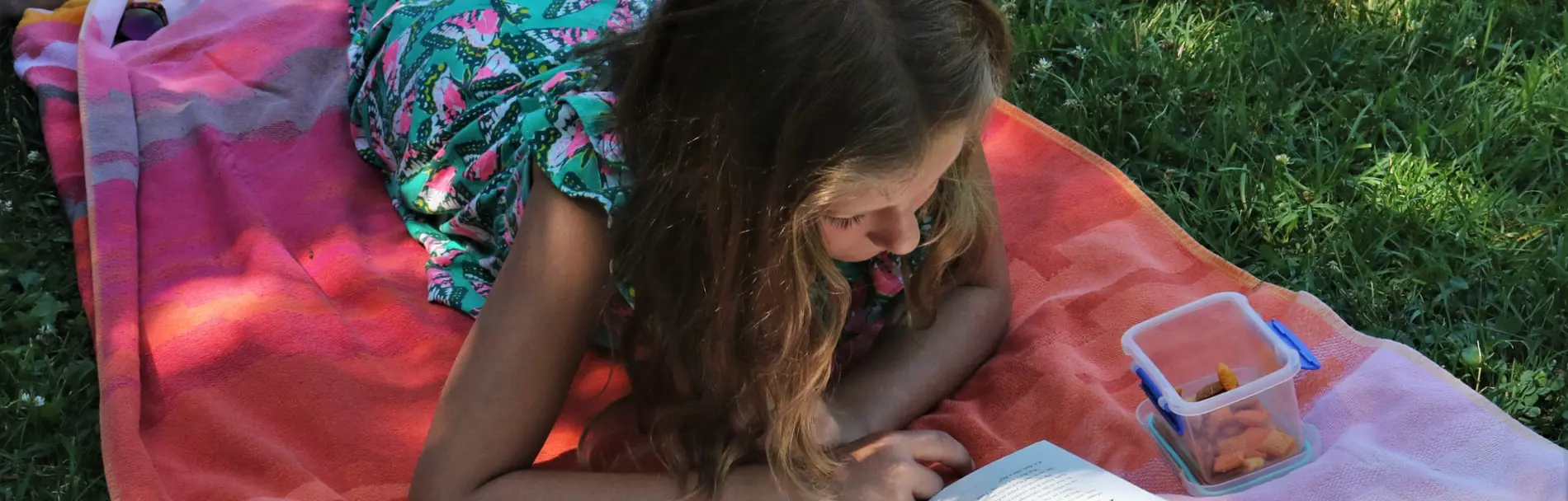 Flicka ligger på en filt i gräset och läser en bok