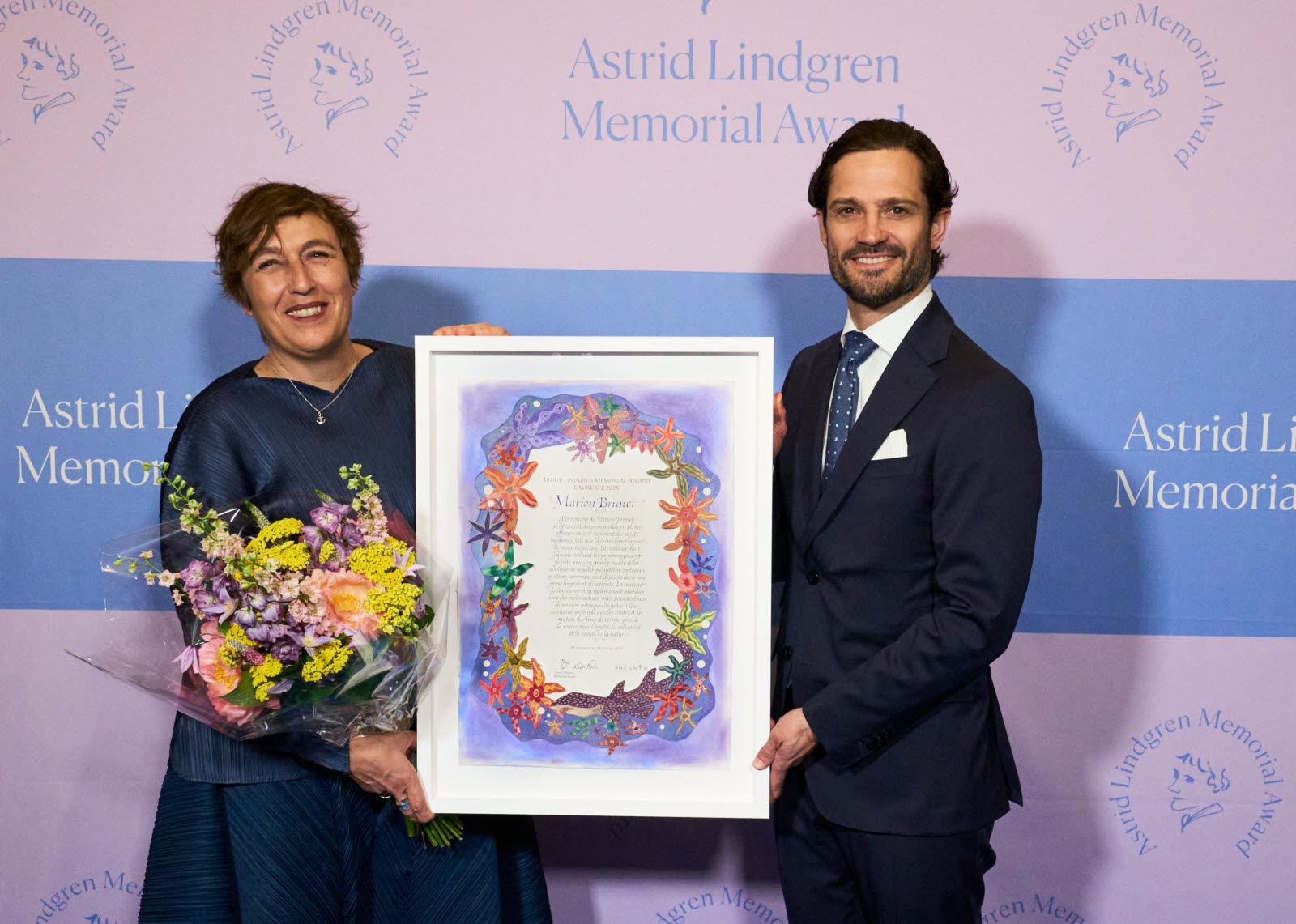 Marion Brunet and Prince Carl Philip holding the award diploma and smiling in front of the Astrid Lindgren Memorial Award logo